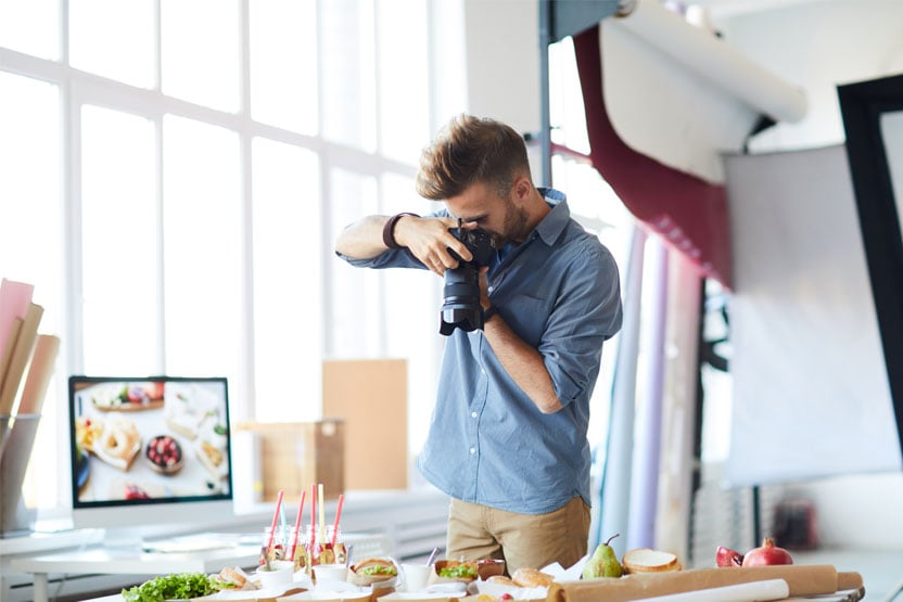 A man holding a camera in a vertical position looking into it whilst photographing items of food on a table.