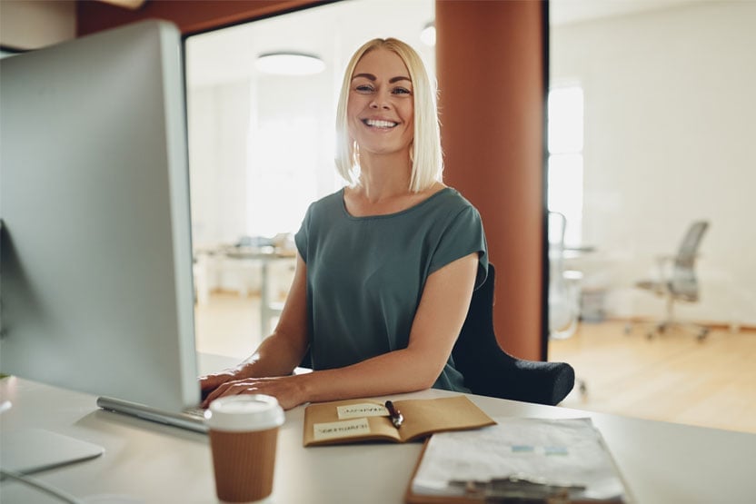 A woman with blonde hair sat in front of computer, smiling at the camera.