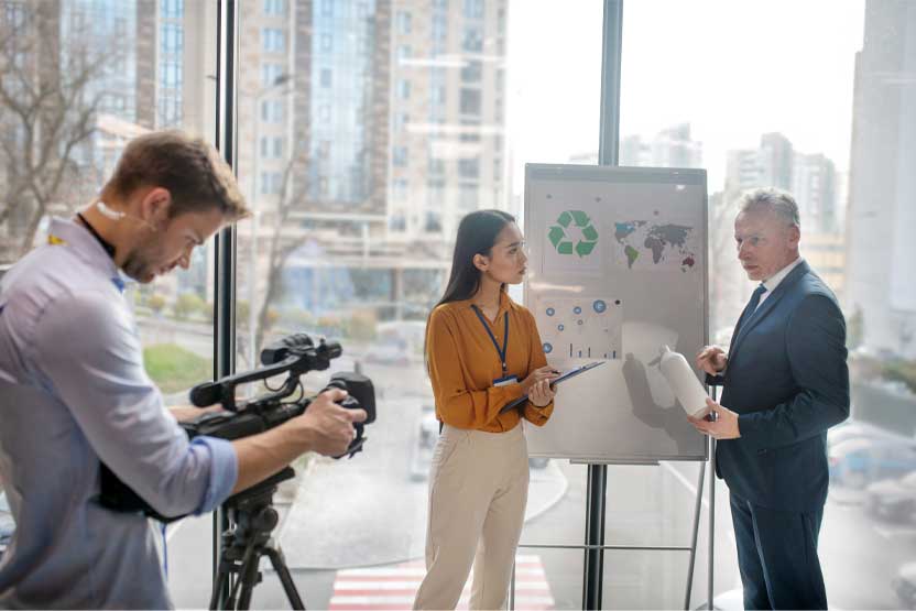 A man looking down at a video camera on a tripod whilst two people are talking in front of a whiteboard.